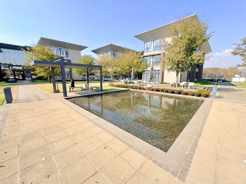 A well-maintained office park courtyard featuring a central water feature, paved walkways, and a modern pergola structure surrounded by contemporary office buildings.