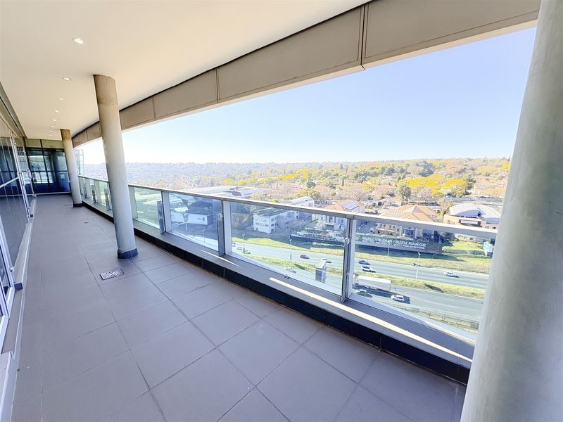 A spacious, modern covered balcony featuring a glass balustrade, tiled flooring, and expansive elevated views of the surrounding area.