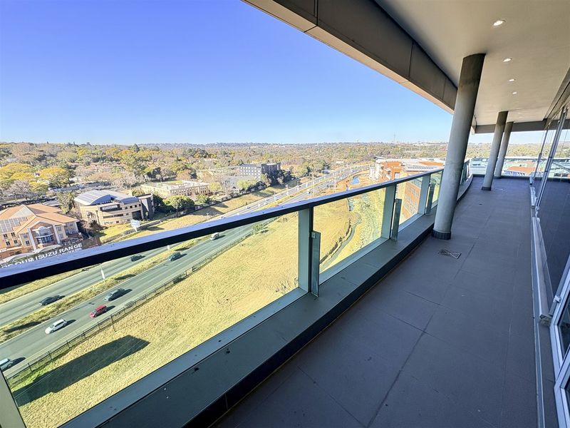 A spacious, modern balcony featuring a glass balustrade and recessed lighting, offering expansive elevated views over the surrounding urban landscape and highway.