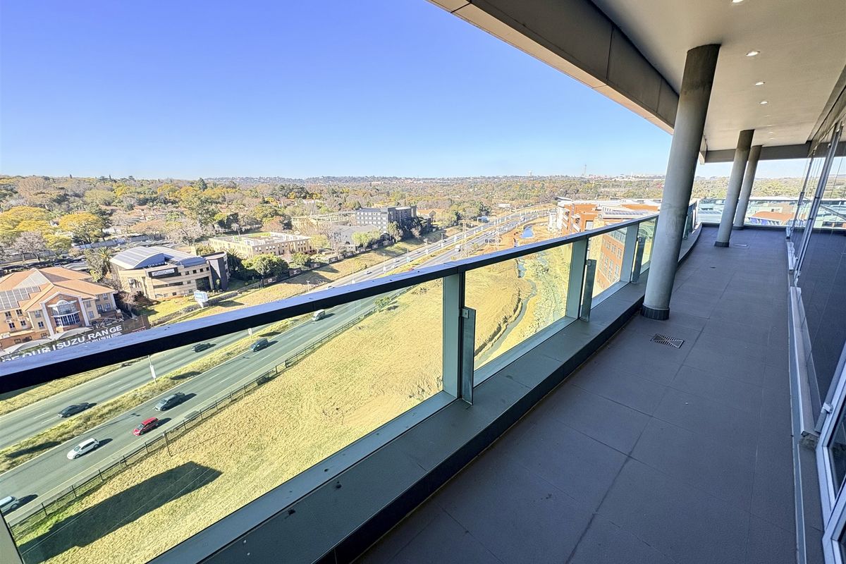 A spacious, modern balcony featuring a glass balustrade and recessed lighting, offering expansive elevated views over the surrounding urban landscape and highway.