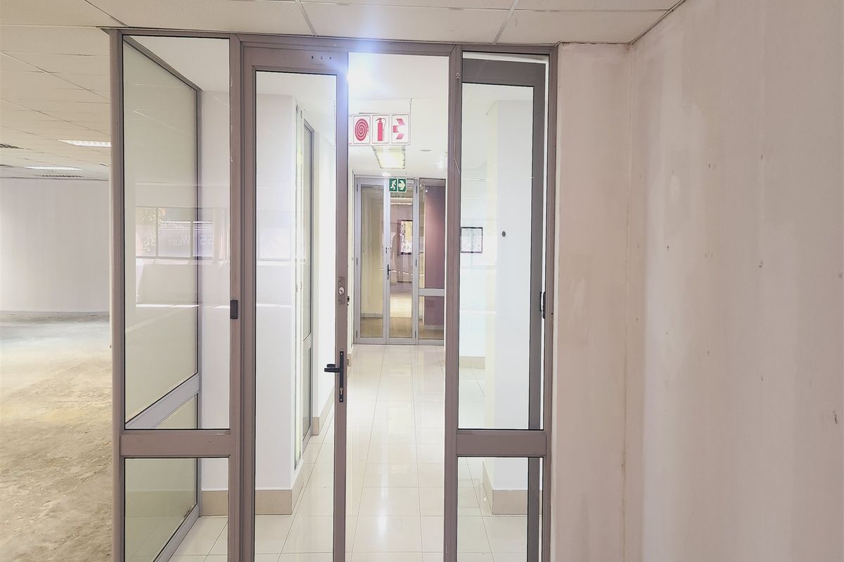 A bright, vacant office hallway featuring tiled flooring, suspended ceilings, and aluminium-framed glass partitioning doors leading to further office spaces.