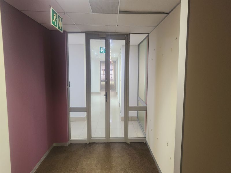 A commercial office hallway featuring a glass partition door, suspended ceiling, and emergency signage, showing signs of wear on the walls and flooring.