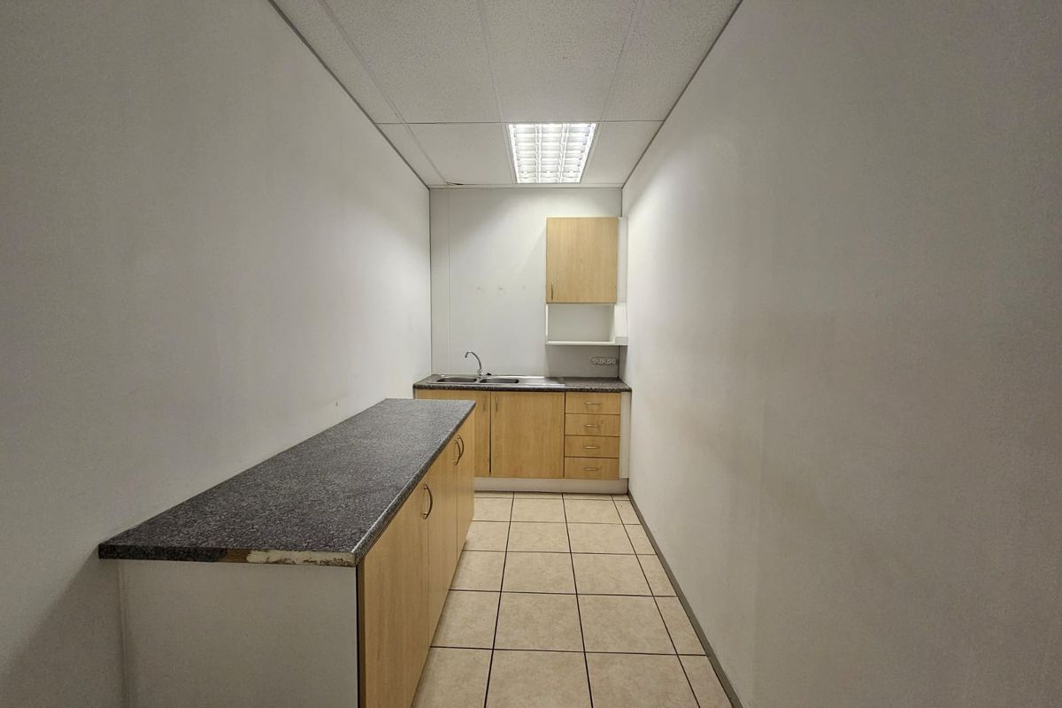 A functional kitchenette featuring light wood cabinetry, laminate countertops, a stainless steel sink, and durable ceramic floor tiles in a clean, empty space.
