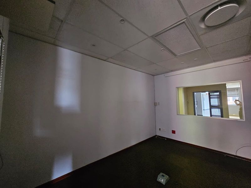 A vacant office space featuring a suspended ceiling, recessed lighting, carpeted flooring, and an internal glass partition providing visibility into the adjacent area.