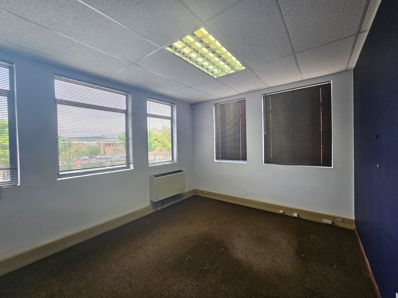 Empty office space featuring a suspended ceiling, recessed lighting, wall-mounted air conditioning unit, and large windows with venetian blinds.