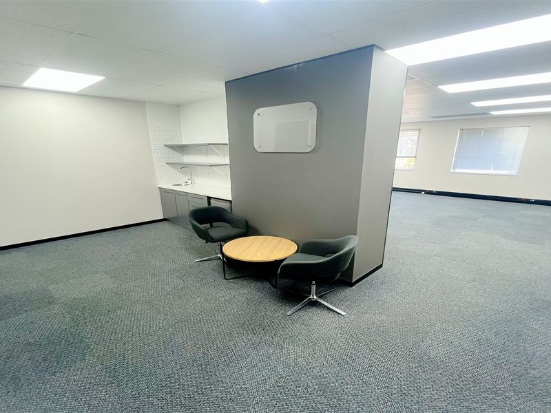 A modern open-plan office space featuring a functional kitchenette, neutral finishes, carpeted flooring, and ample natural light through large windows.