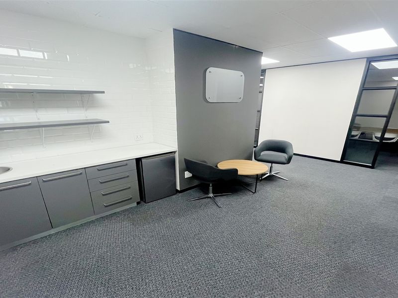 Modern office space featuring a built-in kitchenette with sink, floating shelving, grey cabinetry, and a glass-partitioned meeting area with carpeted flooring.