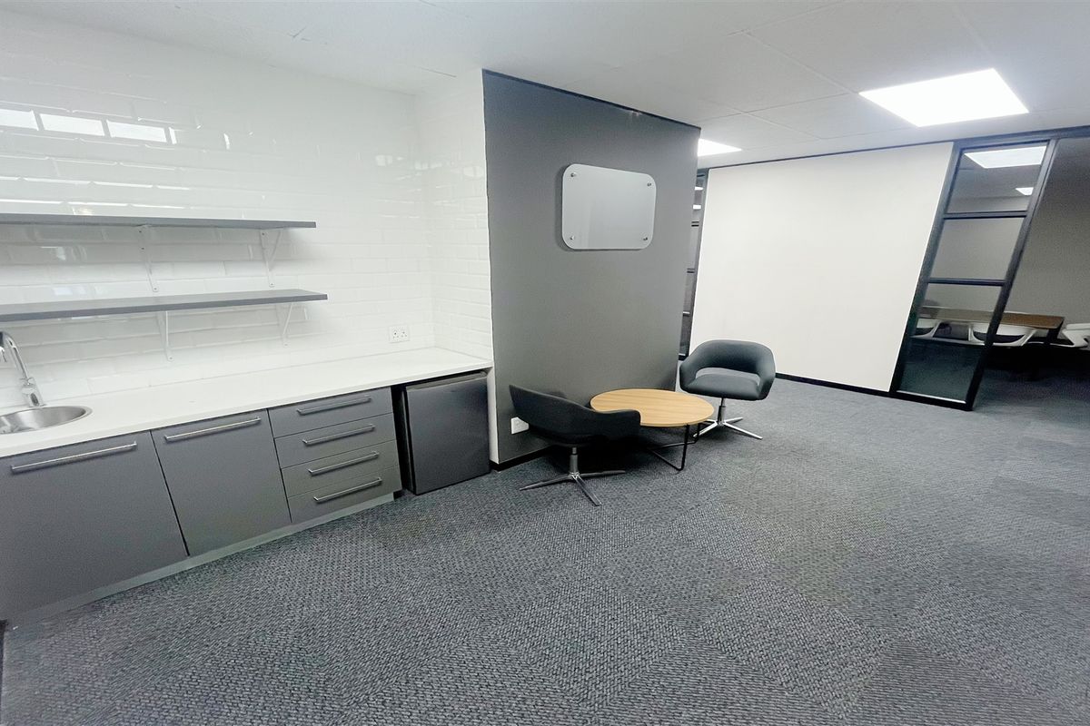 Modern office space featuring a built-in kitchenette with sink, floating shelving, grey cabinetry, and a glass-partitioned meeting area with carpeted flooring.