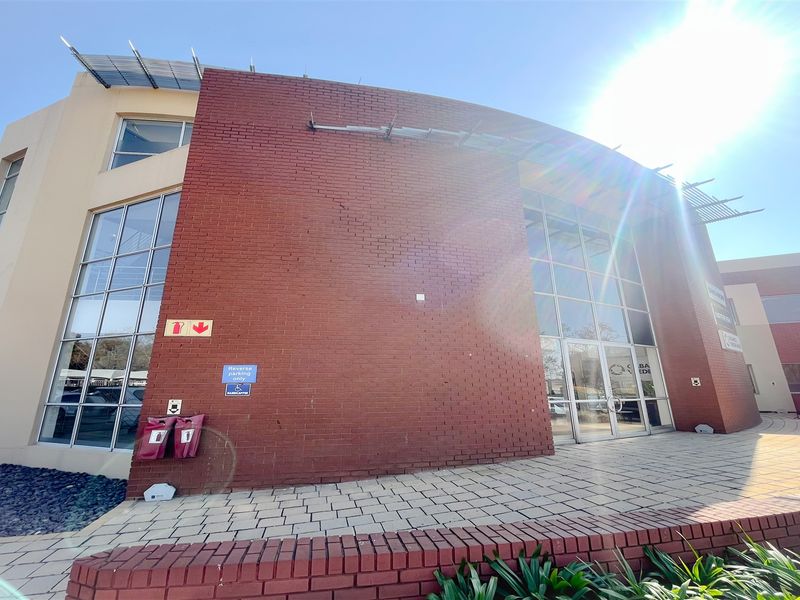 Front elevation of a modern office building featuring a prominent red brick facade, large glass windows, and a paved entrance walkway.
