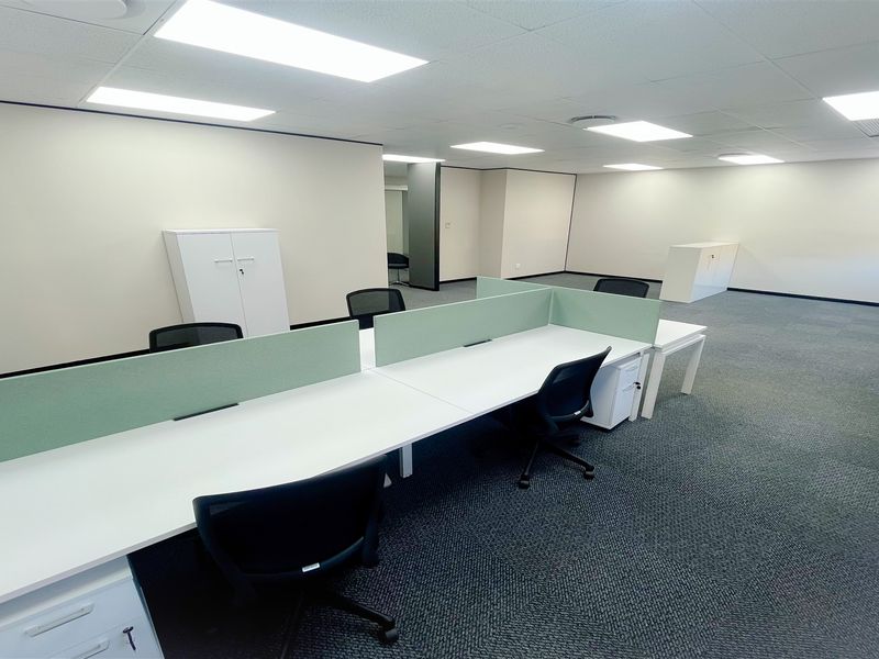 A spacious, well-lit open-plan office area featuring neutral walls, carpeted flooring, and integrated ceiling lighting, suitable for a professional workspace.