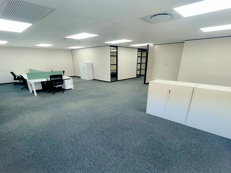 A spacious, well-lit open-plan office area featuring carpeted flooring, suspended ceilings with recessed lighting, and modern glass partition walls.
