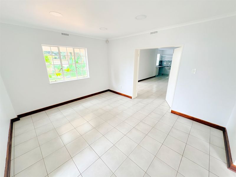 A bright, empty office space featuring neutral tiled flooring, recessed downlights, and timber skirting boards, with a view into an adjacent kitchen area.
