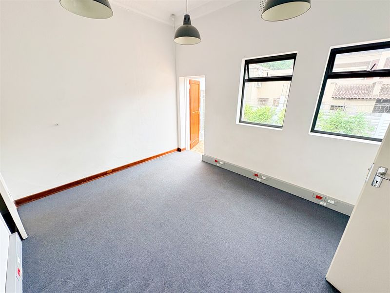 A vacant office space featuring grey carpet flooring, wall-mounted electrical trunking, and natural light from two casement windows.