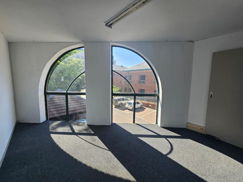 An empty office space featuring distinctive arched windows, grey carpeting, and overhead fluorescent lighting, offering a bright and functional workspace.