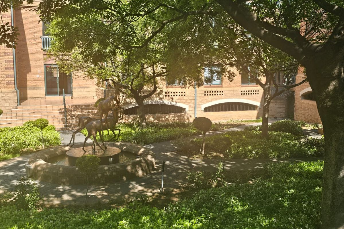 A well-maintained courtyard garden featuring a central stone fountain, mature trees, and paved pathways set against a classic brick building facade.