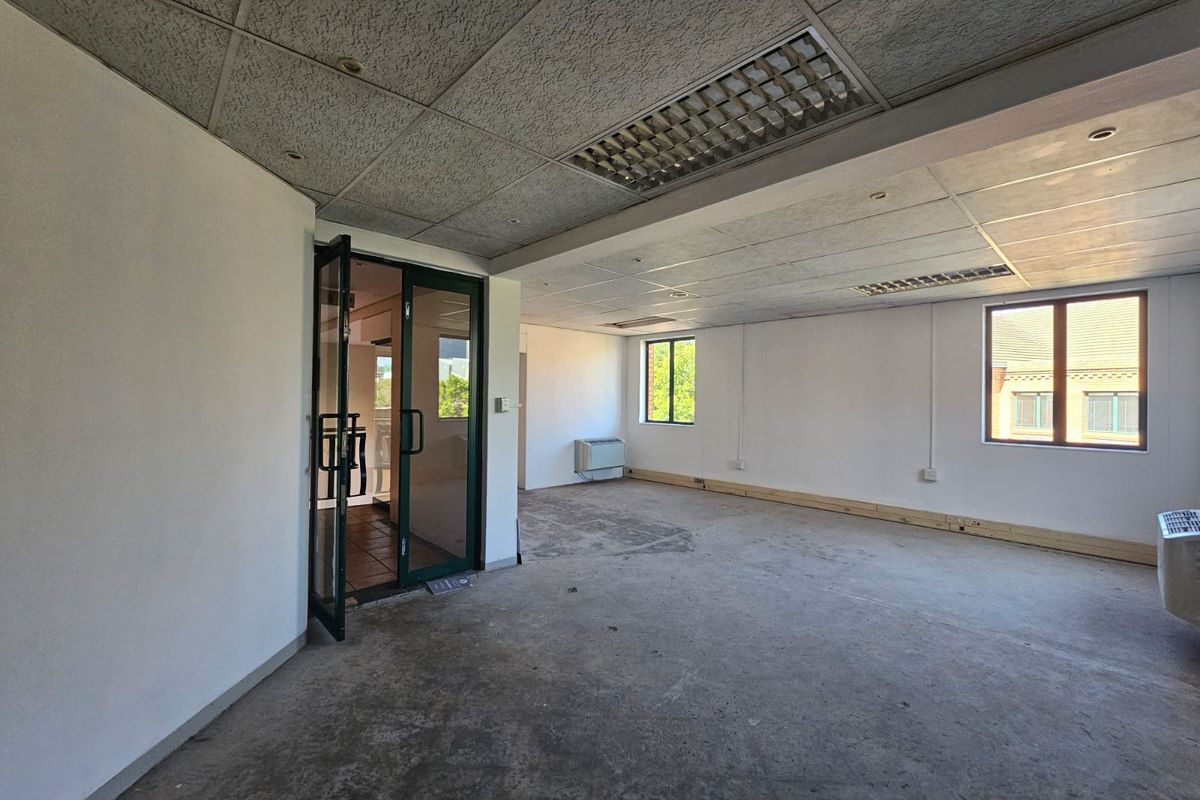 An empty, open-plan office space featuring a suspended ceiling, recessed lighting, air conditioning unit, and large windows providing natural light.