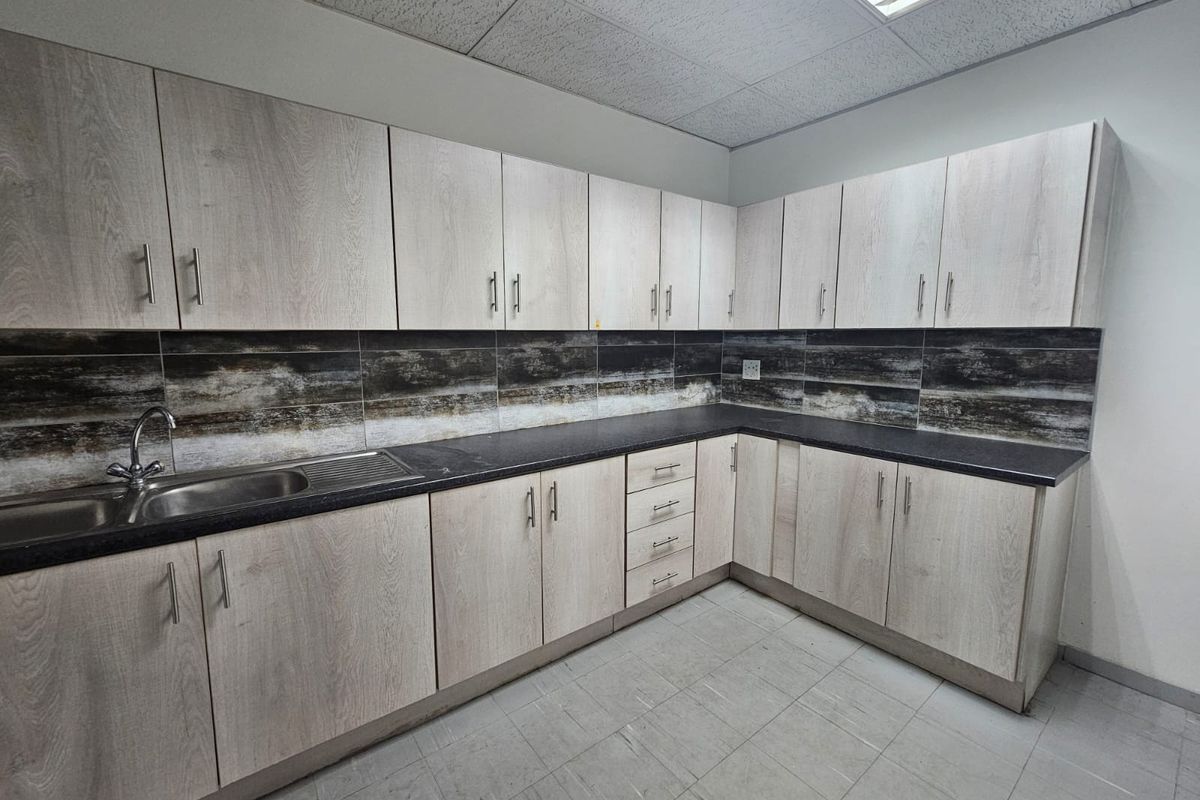 A clean, functional kitchenette featuring light wood-grain cabinetry, dark countertops, a stainless steel sink, and a decorative tiled backsplash.