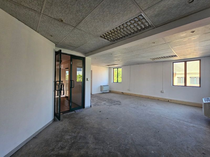 An empty, open-plan office space featuring a suspended ceiling, recessed lighting, air conditioning unit, and large windows providing natural light.