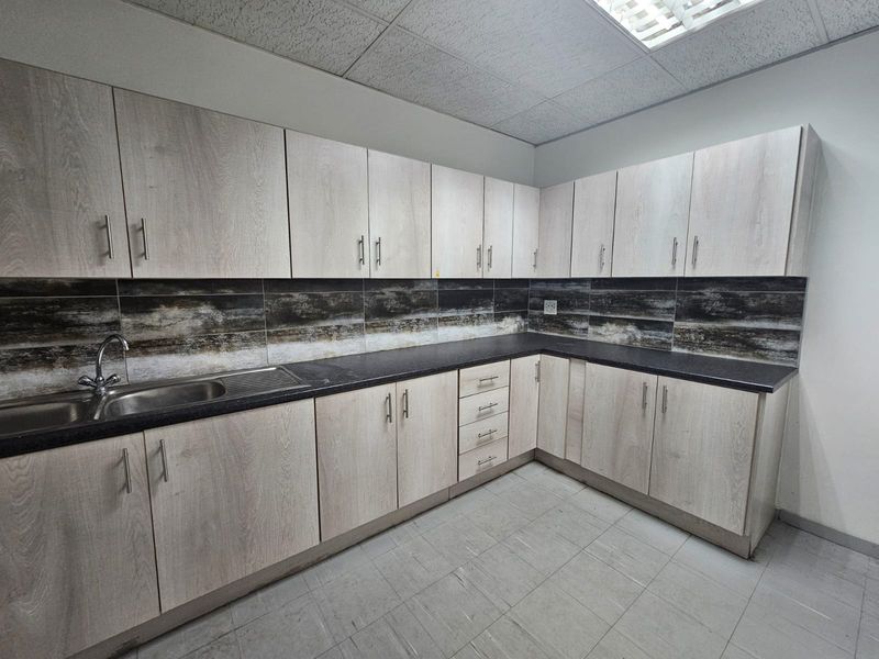 A clean, functional kitchenette featuring light wood-grain cabinetry, dark countertops, a stainless steel sink, and a decorative tiled backsplash.