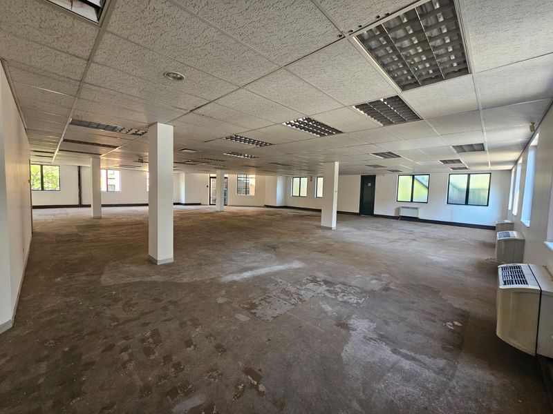 A large, open-plan office space featuring a suspended ceiling, structural columns, and wall-mounted air conditioning units, currently in need of flooring and ceiling refurbishment.