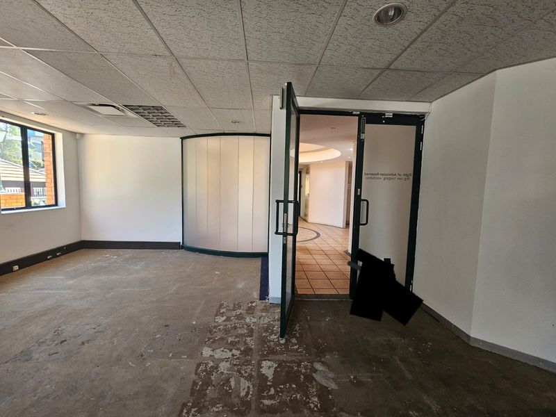 Empty office space featuring a suspended ceiling, recessed lighting, a curved frosted glass partition, and double glass entrance doors leading to a tiled hallway.