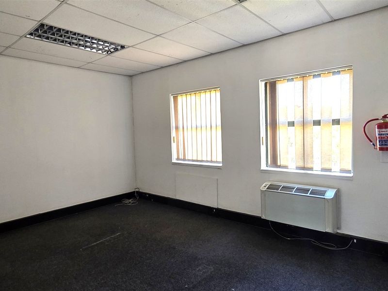 A vacant office space featuring carpet flooring, a suspended ceiling with recessed lighting, vertical blinds, and a wall-mounted HVAC unit.