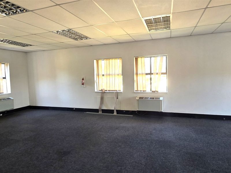 A spacious, empty office area featuring suspended ceilings, recessed lighting, carpeted floors, and wall-mounted air conditioning units.