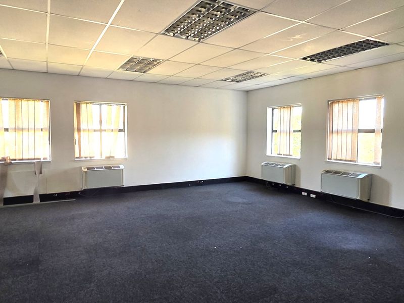 A spacious, empty office area featuring carpeted flooring, suspended ceilings with recessed lighting, and wall-mounted HVAC units for climate control.