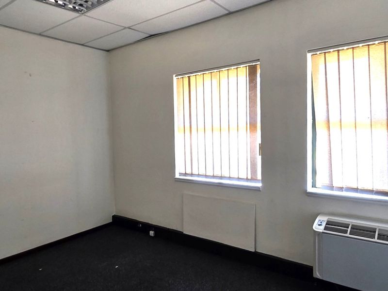 An empty office space featuring a suspended ceiling, carpeted flooring, vertical blinds, and a wall-mounted HVAC unit.