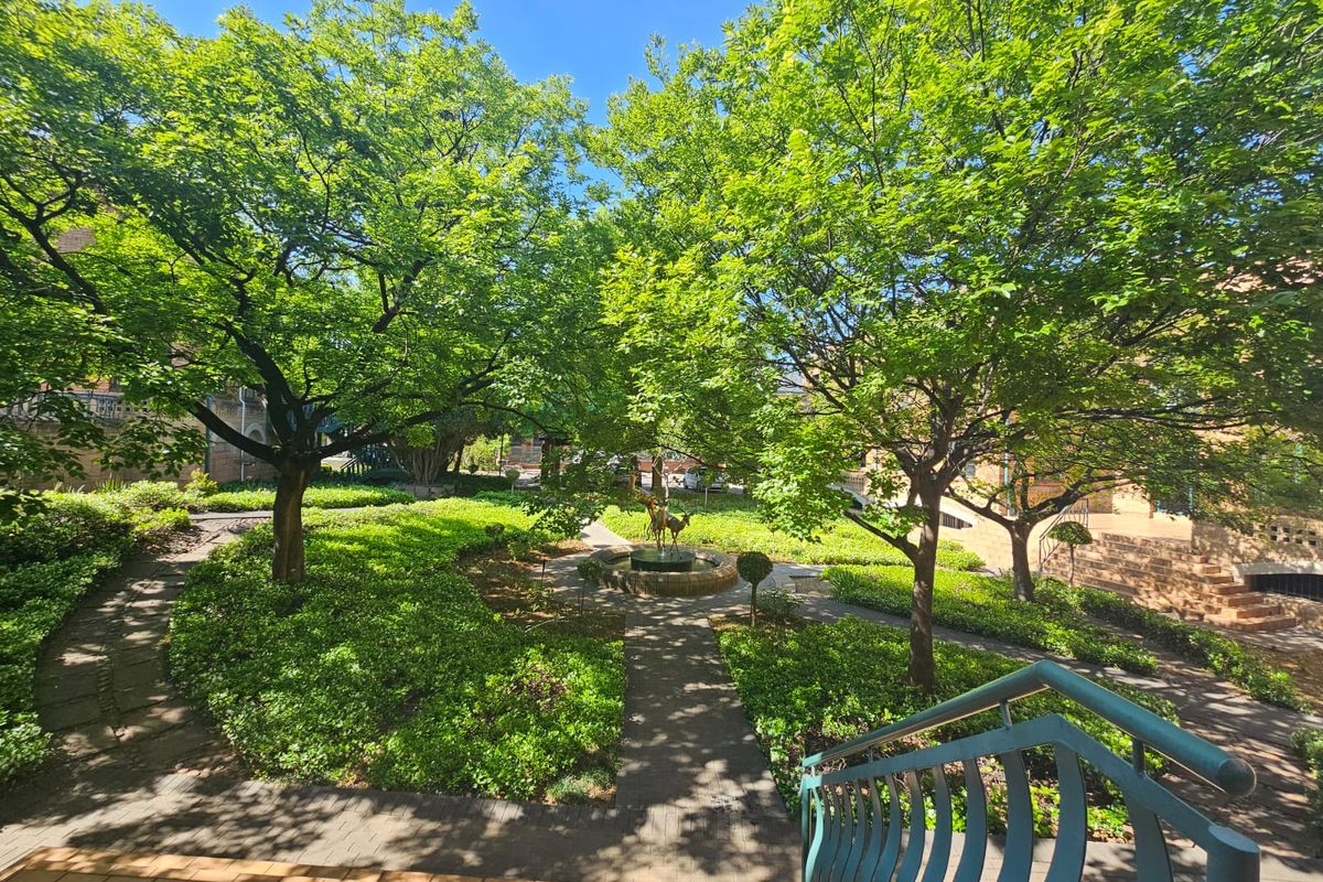 A well-maintained landscaped garden featuring mature trees, paved walkways, and a central stone fountain, viewed from an elevated staircase.