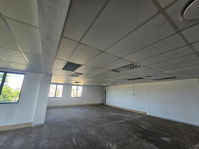 An empty, open-plan office space featuring a suspended ceiling with recessed lighting, air conditioning vents, and large windows providing natural light.