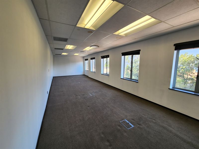 A spacious, empty office area featuring carpeted flooring, recessed lighting, and multiple windows providing natural light.