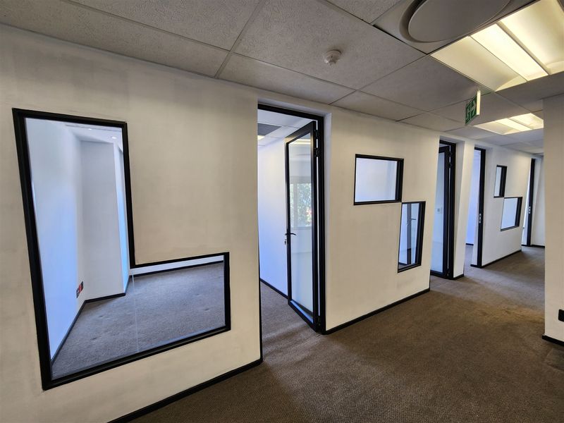 A bright office hallway featuring modern glass partitions, black metal framing, suspended ceilings with recessed lighting, and neutral carpeting throughout the workspace.