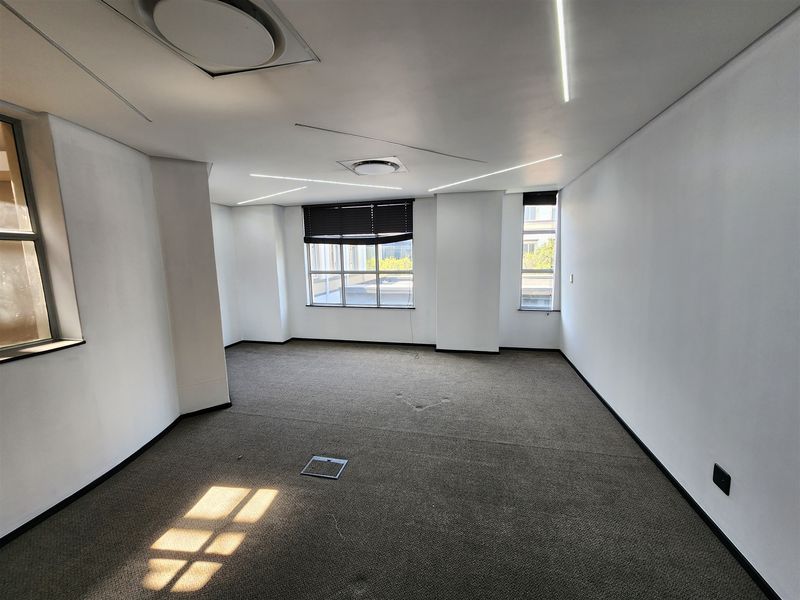 A vacant, well-lit office space featuring neutral carpeted flooring, recessed ceiling lighting, and large windows with fitted blinds.