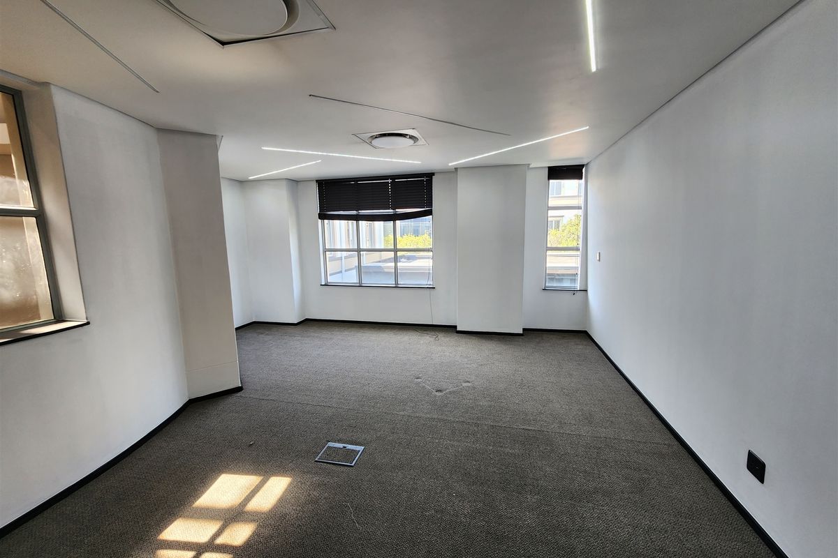 A vacant, well-lit office space featuring neutral carpeted flooring, recessed ceiling lighting, and large windows with fitted blinds.