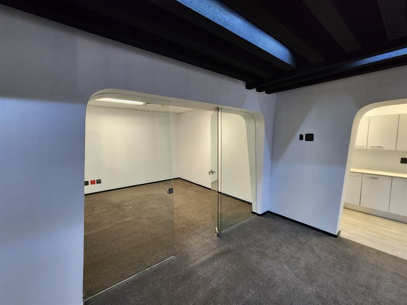 A modern office space featuring a glass partition wall, carpeted flooring, and access to a small kitchenette area with built-in cabinetry.