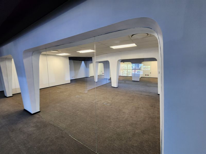 A spacious, vacant open-plan office area featuring modern glass partitions, suspended ceilings with recessed lighting, and carpeted flooring throughout.