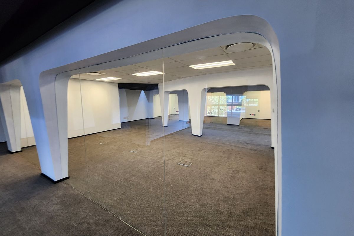A spacious, vacant open-plan office area featuring modern glass partitions, suspended ceilings with recessed lighting, and carpeted flooring throughout.
