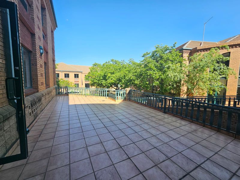 A spacious, tiled outdoor balcony featuring a metal balustrade and views of the surrounding brick office complex and mature trees.