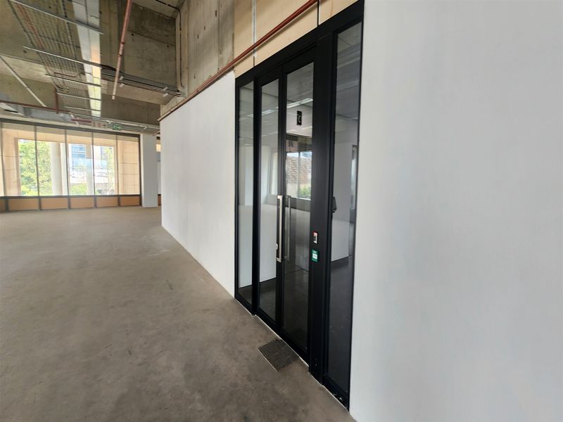 An open-plan office space featuring an industrial aesthetic with exposed concrete ceilings, large windows, and a modern glass-partitioned entrance.