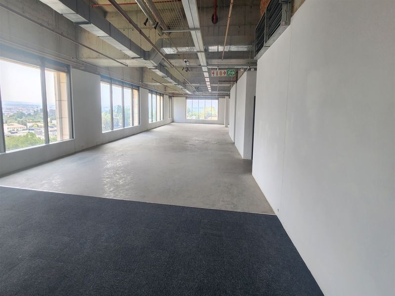An expansive, open-plan office space featuring exposed industrial ceilings, large windows with natural light, and a mix of carpet and concrete flooring.