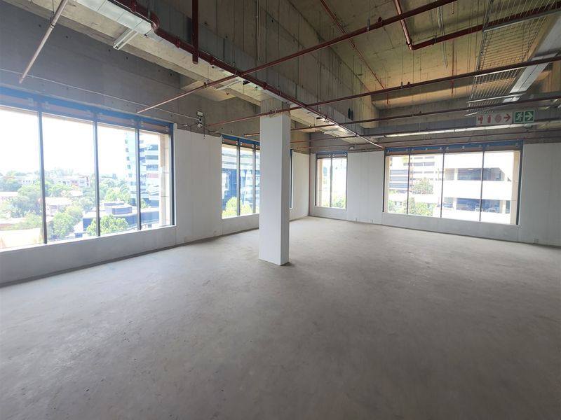 A spacious, open-plan office shell featuring exposed industrial ceilings, concrete flooring, and expansive windows providing ample natural light and urban views.