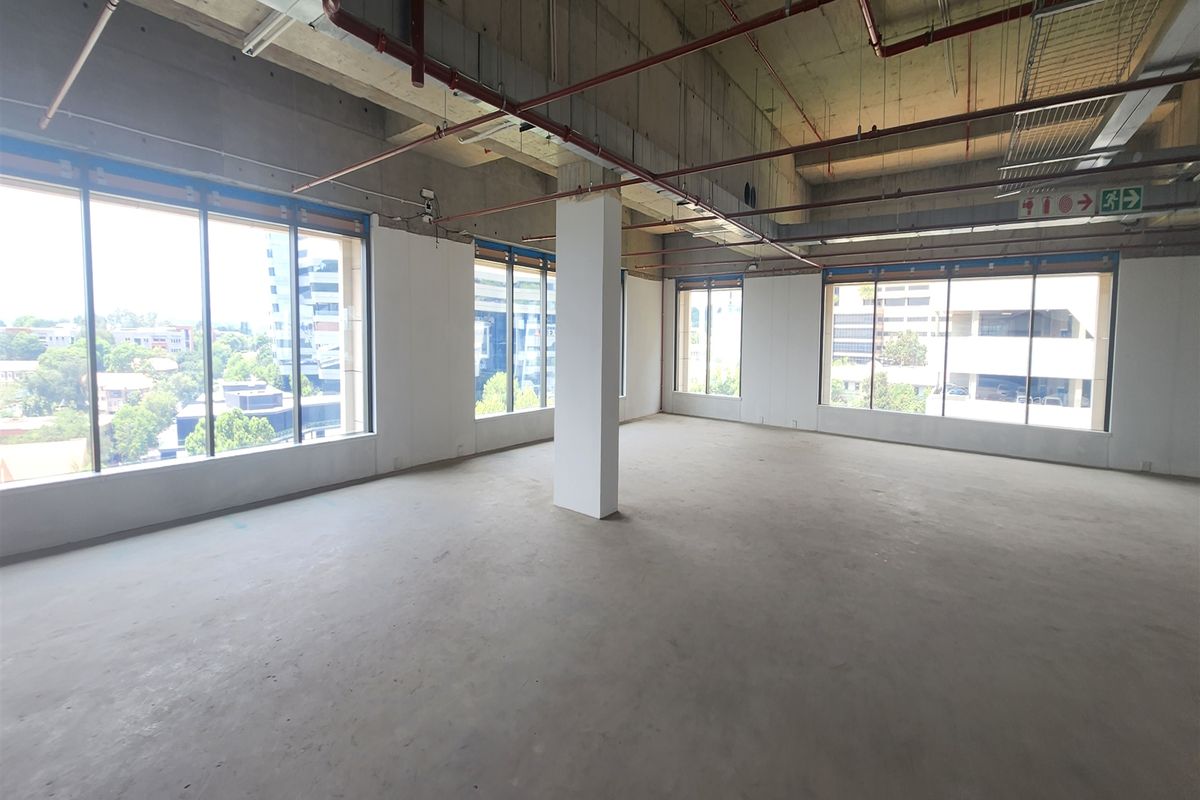A spacious, open-plan office shell featuring exposed industrial ceilings, concrete flooring, and expansive windows providing ample natural light and urban views.