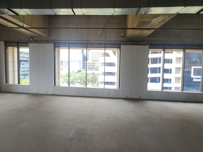 An open-plan office space in shell condition featuring exposed industrial ceilings, concrete flooring, and large windows providing ample natural light and city views.