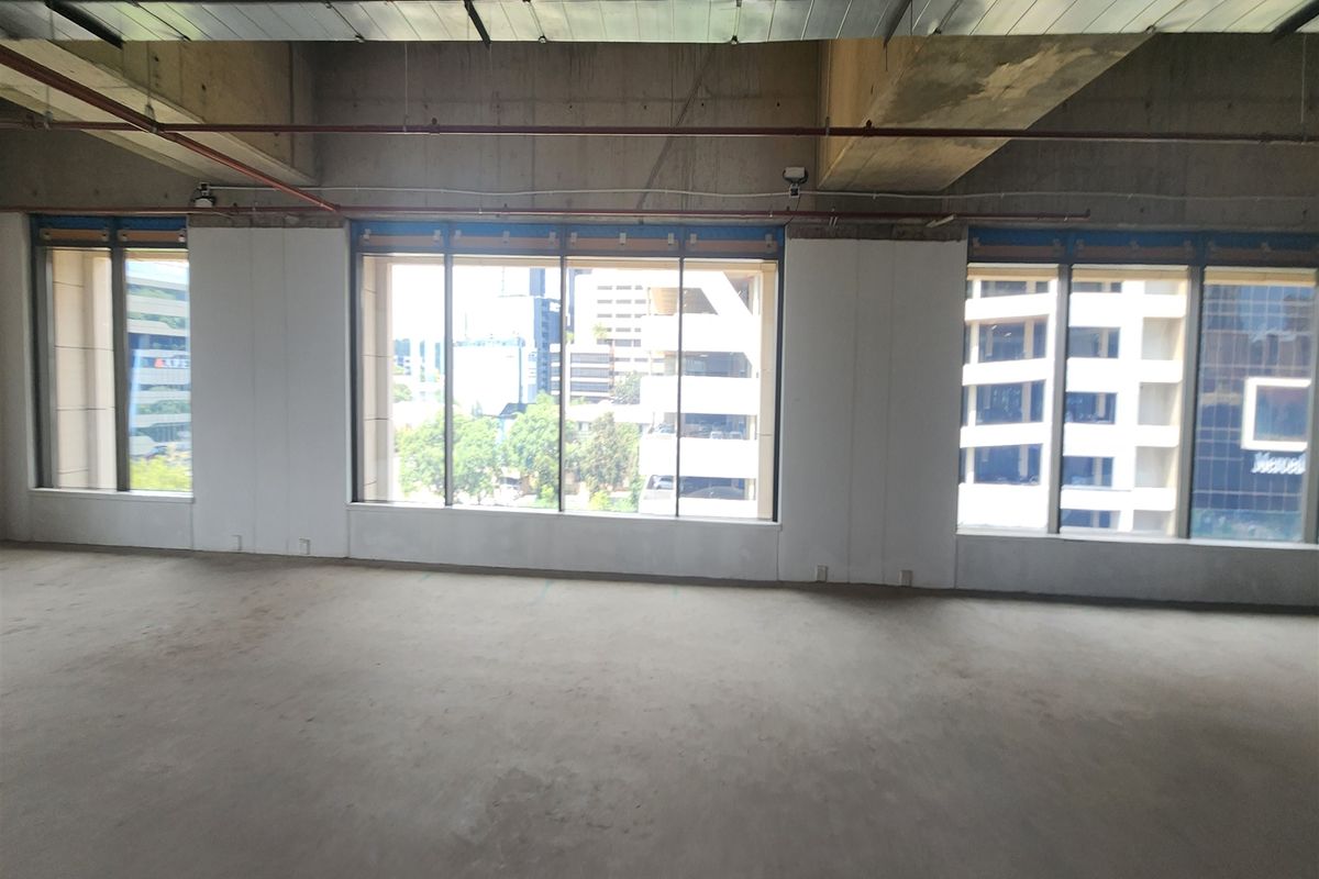 An open-plan office space in shell condition featuring exposed industrial ceilings, concrete flooring, and large windows providing ample natural light and city views.