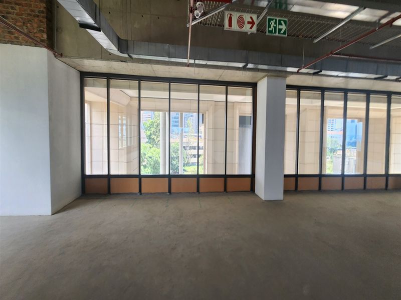 An open-plan office space featuring exposed concrete ceilings, industrial ducting, and large floor-to-ceiling windows providing ample natural light and city views.