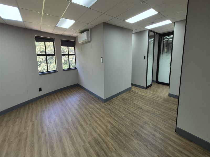 A modern, vacant office space featuring wood-look flooring, recessed ceiling lights, a split-system air conditioner, and a glass-panelled entrance door.