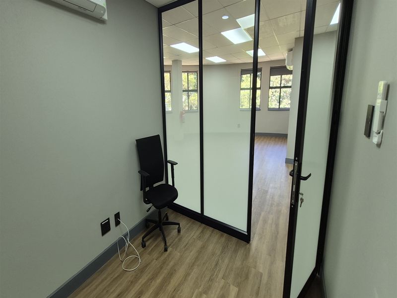 A modern office space featuring a glass partition wall, suspended ceiling with recessed lighting, and wood-look flooring, offering a clean and professional work environment.