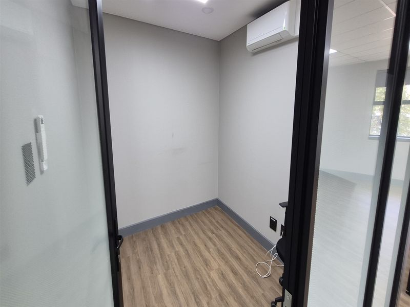 A compact, empty office space featuring modern glass partitioning, wood-look laminate flooring, and a wall-mounted air conditioning unit.