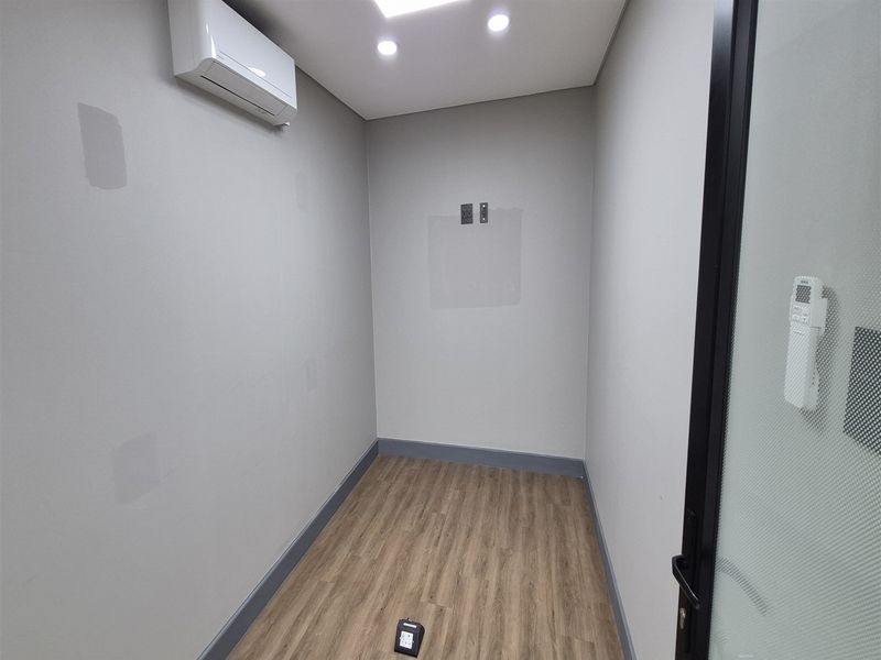 A compact, empty office space featuring laminate flooring, recessed lighting, a wall-mounted air conditioning unit, and glass-panelled entry door.